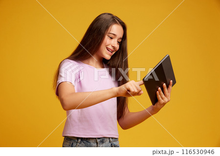 Happy smiling teen girl with brown long hair, in pink t-shirt, using tablet for leisure and education against yellow studio background 116530946
