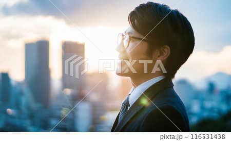 ビル群を背景に微笑む若いビジネスマンの横顔のアップ。スーツを着てメガネをかけた若い男性。 116531438