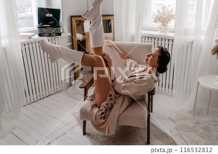 A woman is sitting in a chair with her legs spread apart. She is wearing white boots and a white dress. The room is decorated with a white color theme. 116532812