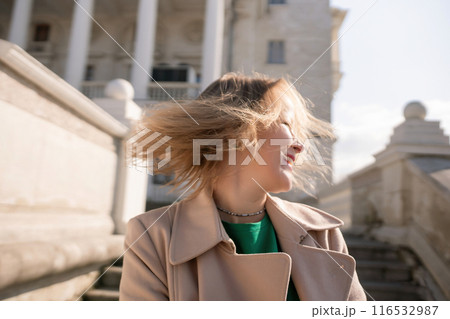 A woman with long hair is standing on a set of stairs. She is wearing a green shirt and a tan coat. A woman with long hair is standing on a set of stairs. She is wearing a green shirt and a tan coat. 116532987