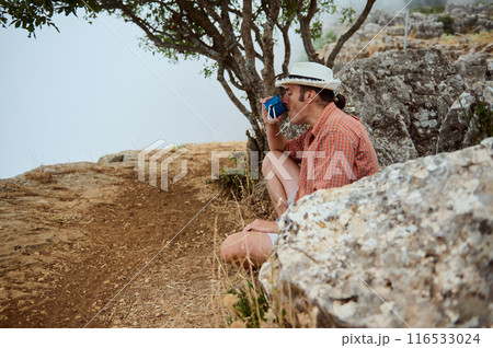 Relaxing hiker enjoying a break with a cup of coffee in nature, sitting by a tree in a tranquil outdoor setting Relaxing hiker enjoying a break with a cup of coffee in nature, sitting by a tree in a tranquil outdoor setting 116533024