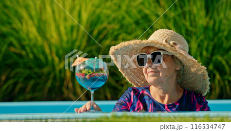 Elderly woman with a delighted expression, wearing a sun hat and sipping a cold drink while lounging in a pool on a bright day 116534747