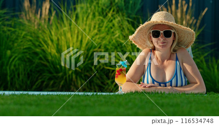 Attractive middle-aged woman in a hat with a cocktail, peeking out of a pool 116534748