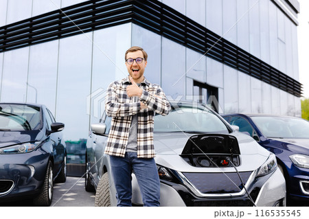 A young man stands next to his new electric car and rests while the car is charging 116535545