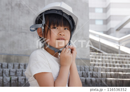 白いヘルメットをかぶった小学生の女の子はコンクリート建物の高台にいる 116536352