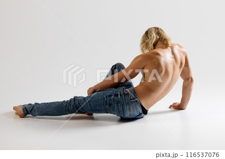 Rear view photo of young athletic man posing sitting in jeans on floor against grey studio background. Rear view photo of young athletic man posing sitting in jeans on floor against grey studio background. 116537076