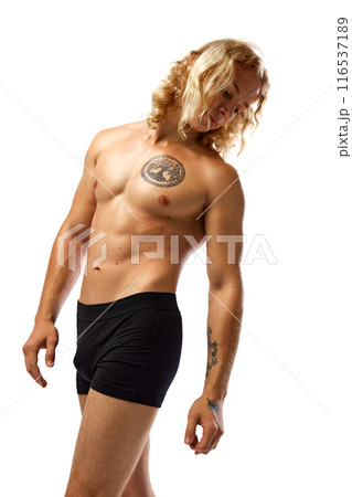 Muscular man with long, curly blonde hair stands in black boxer briefs, showing off his chest tattoo and arm tattoos. against white studio background. Muscular man with long, curly blonde hair stands in black boxer briefs, showing off his chest tattoo and arm tattoos. against white studio background. 116537189