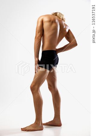 Muscular young man with blonde hair stands in black underwear showing his back against white studio background. 116537191