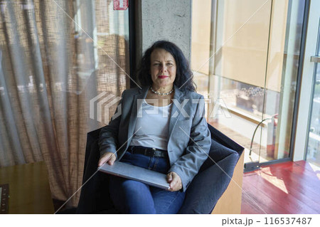 A 57-year-old woman sits with a laptop in a chair in a modern workspace 116537487