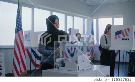 Muslim woman in hijab puts ballot paper in box at polling station 116537655