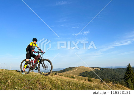 Cyclist man riding electric bike outdoors on sunny day. Male tourist resting on grassy hill, enjoying beautiful mountain landscape, wearing helmet and backpack. Concept of active leisure. Cyclist man riding electric bike outdoors on sunny day. Male tourist resting on grassy hill, enjoying beautiful mountain landscape, wearing helmet and backpack. Concept of active leisure. 116538181