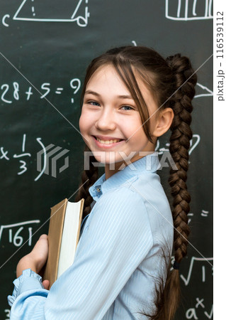 A high school schoolgirl stands near the blackboard with a textbook in her hands, portrait of a teenage girl at school 116539112