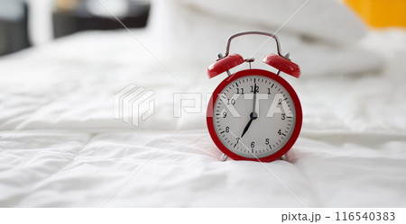 Red alarm clock at 7 oclock on bed 116540383