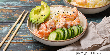 White ceramic bowl with rice, shrimps, avocado, vegetables and sesame seeds and chopsticks on colourful rustic wooden background front view. Healthy asian style poke bowl White ceramic bowl with rice, shrimps, avocado, vegetables and sesame seeds and chopsticks on colourful rustic wooden background front view. Healthy asian style poke bowl 116542586