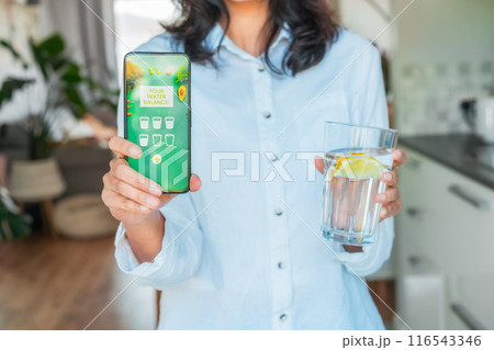 Healthy habit to drink water. Blurred no face woman with glass of pure water with lemon holding phone with active mobile app to control body hydration, Tracking water balance. Selective focus. 116543346
