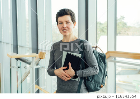 Smiling neutral gender middle-aged female person in casual clothes with backpack and books stands on staircase in modern building of university or education Centre. Adult students, Academic education. 116543354