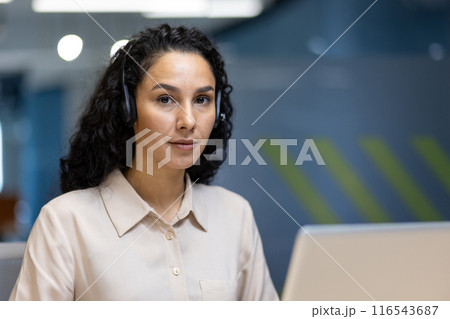 Focused professional woman with curly hair and headset working at desk in office environment. Modern workplace with blue and green decor. Ideal image depicting customer service, support, and 116543687