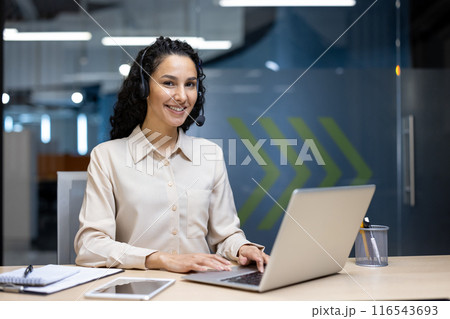 Happy customer service representative wearing headset working on laptop in modern office environment. Professional setting depicting workplace and technology. 116543693
