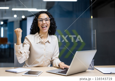 Thrilled businesswoman in glasses celebrates a professional achievement in a modern office. She is seen seated at a desk with a laptop, tablet, and smartphone, expressing success. 116543719