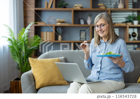 Senior woman eating salad while working from home on laptop. Comfortable home office setup with modern interior. Retirement, remote work, and healthy living concepts. 116543728