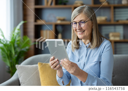 Smiling senior woman using digital tablet at home, sitting on sofa. Enjoying technology, staying connected with family and friends. Modern living room with natural light, bookshelf, and green plant. Smiling senior woman using digital tablet at home, sitting on sofa. Enjoying technology, staying connected with family and friends. Modern living room with natural light, bookshelf, and green plant. 116543738