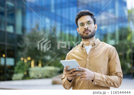 Confident hispanic man wearing glasses and casual clothes using digital tablet outdoors near modern office building. Urban office background with greenery and glass architecture. Confident hispanic man wearing glasses and casual clothes using digital tablet outdoors near modern office building. Urban office background with greenery and glass architecture. 116544444