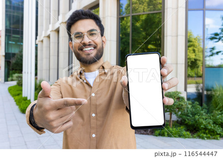 Smiling hispanic man outdoors pointing at blank smartphone screen wearing brown shirt against office building and greenery background Smiling hispanic man outdoors pointing at blank smartphone screen wearing brown shirt against office building and greenery background 116544447