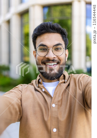 Cheerful hispanic man with glasses taking selfie outside modern office building on sunny day, smiling at camera. Business casual attire, outdoor setting with green background. 116544448