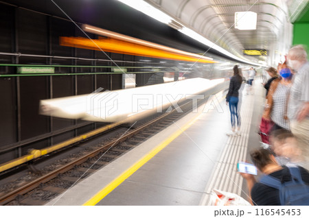 Conceptual image of the subway platform. A trail of light on the tracks evokes the passage of the train, the figures on the platform suggest people waiting with masks, a woman uses her cell phone. 116545453