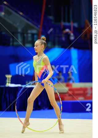 A young rhythmic gymnast shows her hoop skills at a lively competition in an indoor arena 116546823