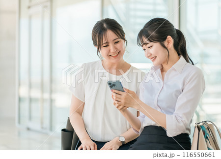 オフィスの休憩所・ベンチでスマホを見ながら笑顔で会話する若いアジア人ビジネスウーマン（同僚・同期） 116550661