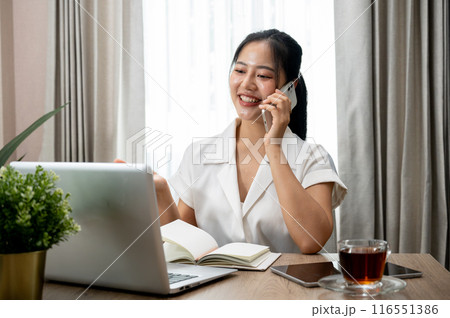 A confident Asian businesswoman is talking on the phone at her desk in her private office. 116551386