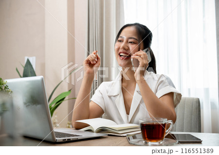 A confident Asian businesswoman is talking on the phone at her desk in her private office. 116551389