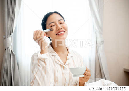 A beautiful, smiling young Asian woman in pajamas is applying a facial treatment mask to her face. 116551455