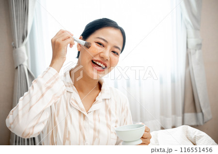 A beautiful, smiling young Asian woman in pajamas is applying a facial treatment mask to her face. 116551456
