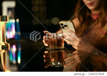 Charming young woman hand holding a glass of whisky using smartphone at bar counter 116557504