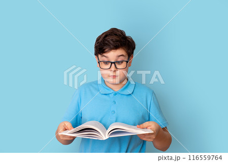 A surprised schoolboy in glasses, holding an open book in his hands, he looks at it A surprised schoolboy in glasses, holding an open book in his hands, he looks at it 116559764
