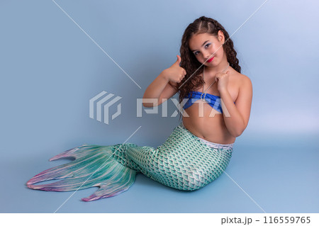Beautiful happy girl in a mermaid costume sitting on a blue background showing thumbs up. Beautiful happy girl in a mermaid costume sitting on a blue background showing thumbs up. 116559765