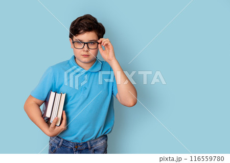 A cute schoolboy in glasses and a blue T-shirt, holding a stack of books A cute schoolboy in glasses and a blue T-shirt, holding a stack of books 116559780