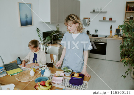 Young Caucasian doctor mom preparing two lunchboxes for her and her kid while her daughter doing homework 116560112
