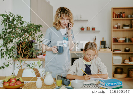 Doctor mother checking daughters homework and speaking on phone while daughter doing exercise in dining area 116560147