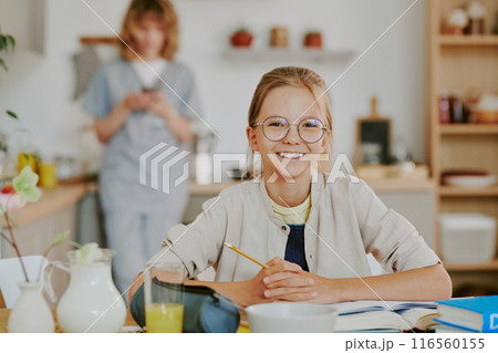Portrait of little girl sitting at table and holding pen while smiling widely, her nurse mom surfing internet in blurred background 116560155