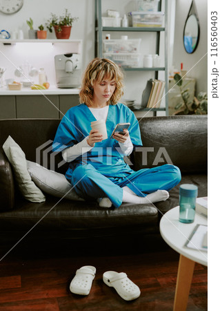 Young Caucasian nurse surfing Internet and holding pastel rose thermal mug with hot coffee while sitting in lotus position on black leather sofa Young Caucasian nurse surfing Internet and holding pastel rose thermal mug with hot coffee while sitting in lotus position on black leather sofa 116560403