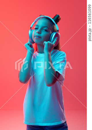 Portrait of young girl, child in casual clothes, white t-shirt listening to music in headphones against pink background in neon light 116561099