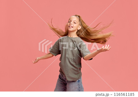 Portrait of young girl with long flowing hair, wearing grey t-shirt and jeans, posing against pink background. Joyful expression and happiness 116561185