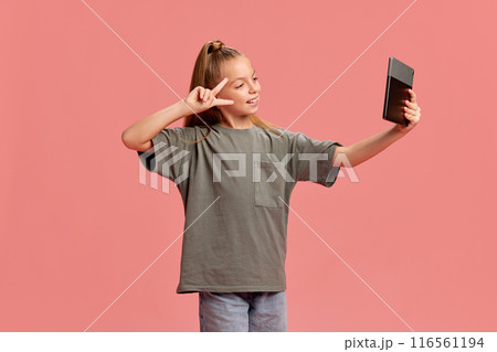 Portrait of young girl in casual clothes taking selfie with tablet, posing on social media, communicating and having fun against peach background 116561194