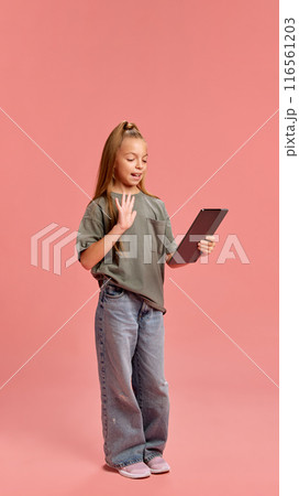Full-length portrait of young girl in jeans and t-shirt using tablet for communication, having online video call against peach background 116561203