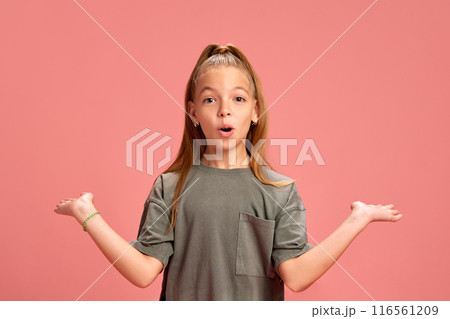 Emotive little girl in casual clothes, grey t0shirt standing with shocked face and spreading hands in surprise against peach background 116561209