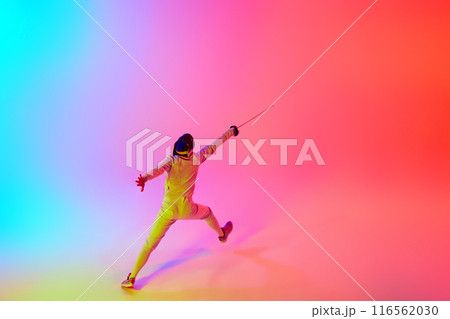 Fencer in gear and mask with their rapier extended forward, performing dynamic leap in mixed neon lights against gradient studio background. 116562030