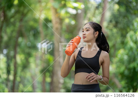 Young Woman Drinking Water from Orange Bottle During Outdoor Workout in Green Park 116564126